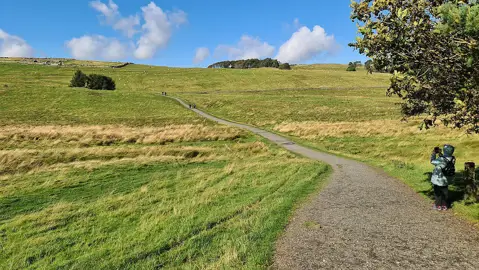 A broad path climbing across grassy fields towards the remains of Housesteads Roman Fort.