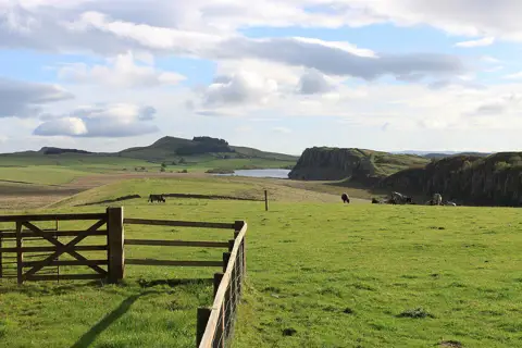 Grassy ground and a gate at Steel Rigg, with crag faces and a lake beyond.